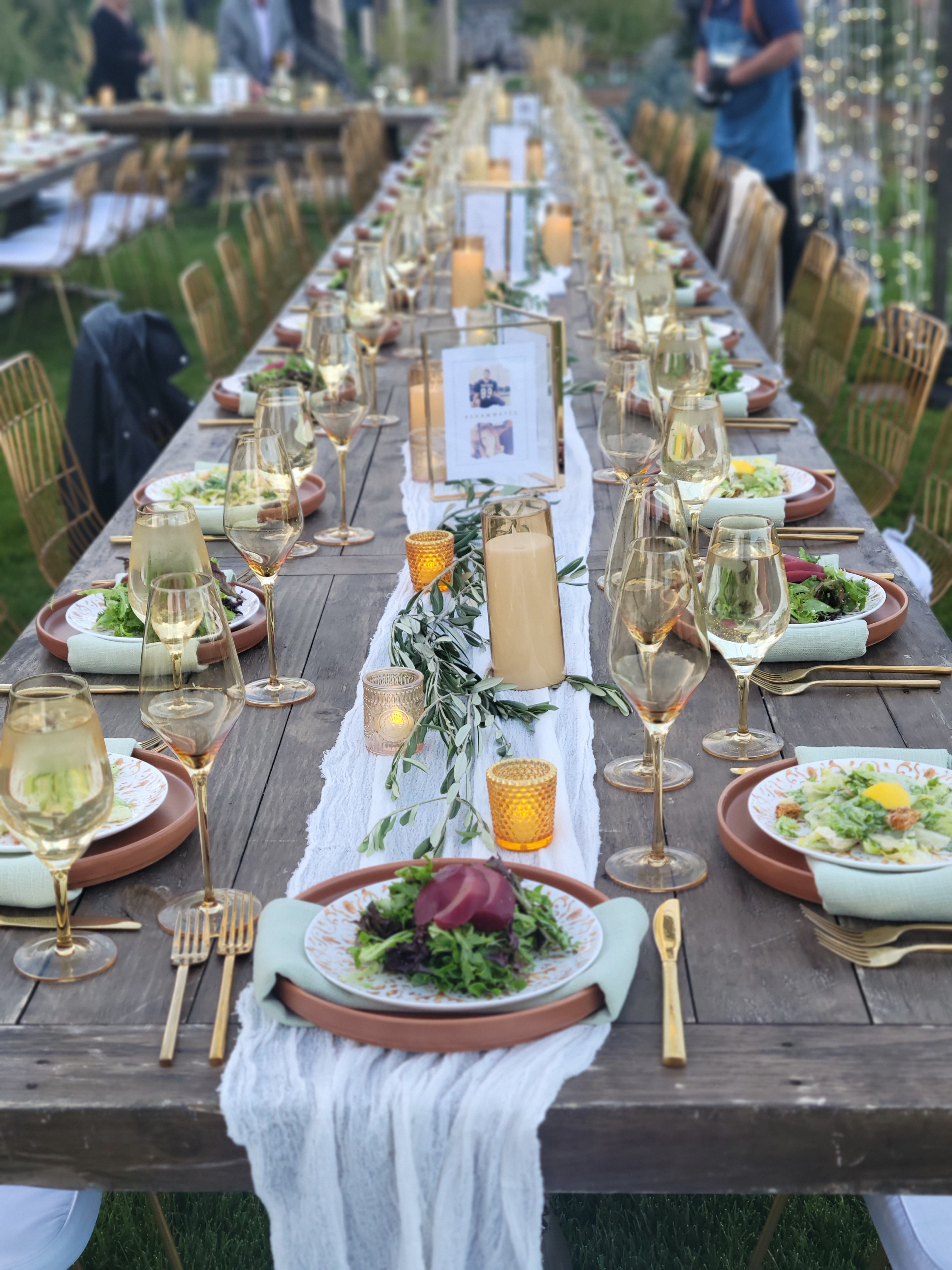 A beautifully set long wooden table outdoors, decorated with elegant dinnerware, candles, and a green centerpiece. Plates of salad and glasses of wine are arranged for a gathering.