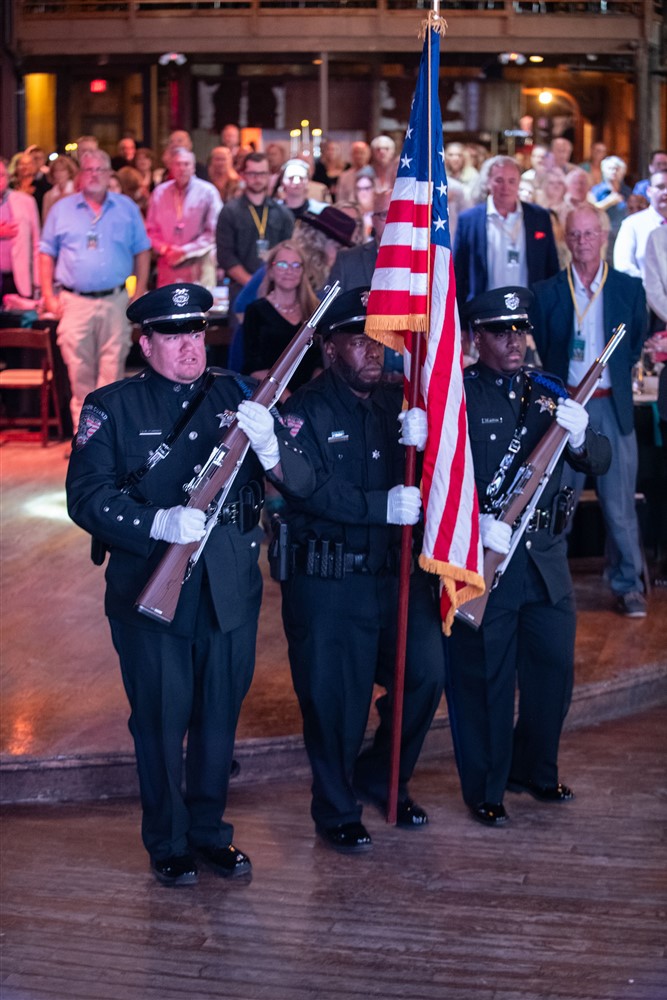 Three uniformed officers in a ceremonial formation, one holding an American flag, while the audience stands in the background.